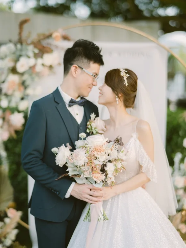 新婚夫婦在淺色花卉拱門前深情相視，手捧米白花束，畫面柔和溫暖。