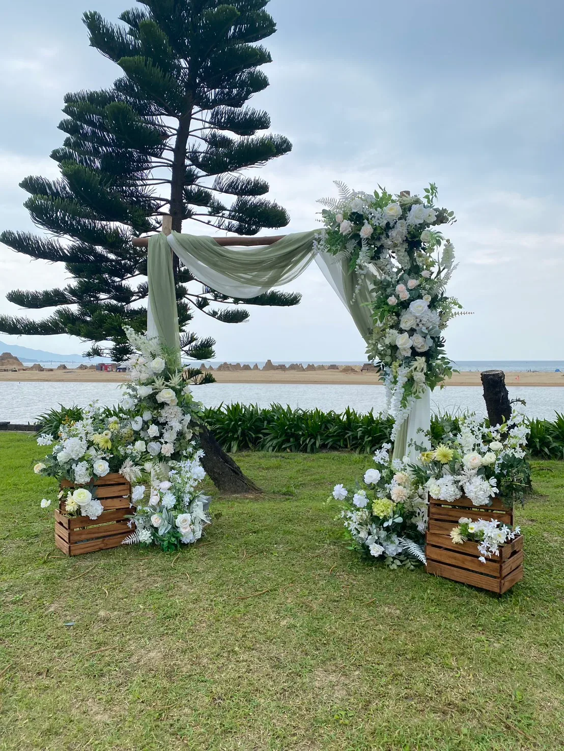 白綠紗幔證婚拱門近照，松樹矗立一側，背景為海景沙灘