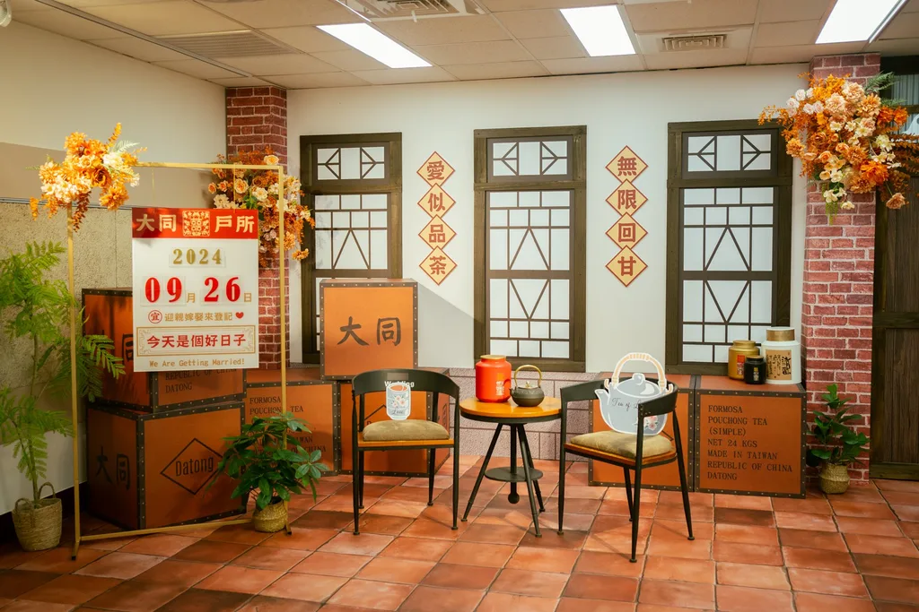 大同戶政事務所結婚登記拍照背板全景,2024 年 9 月施作。實體紅磚柱、三面立體木格窗、老磁磚地,小圓桌上放錫製茶葉罐與鐵壺,牆上菱形牌對聯「愛似品茶/無限回甘」,左側日期牌停在 2024.09.26。