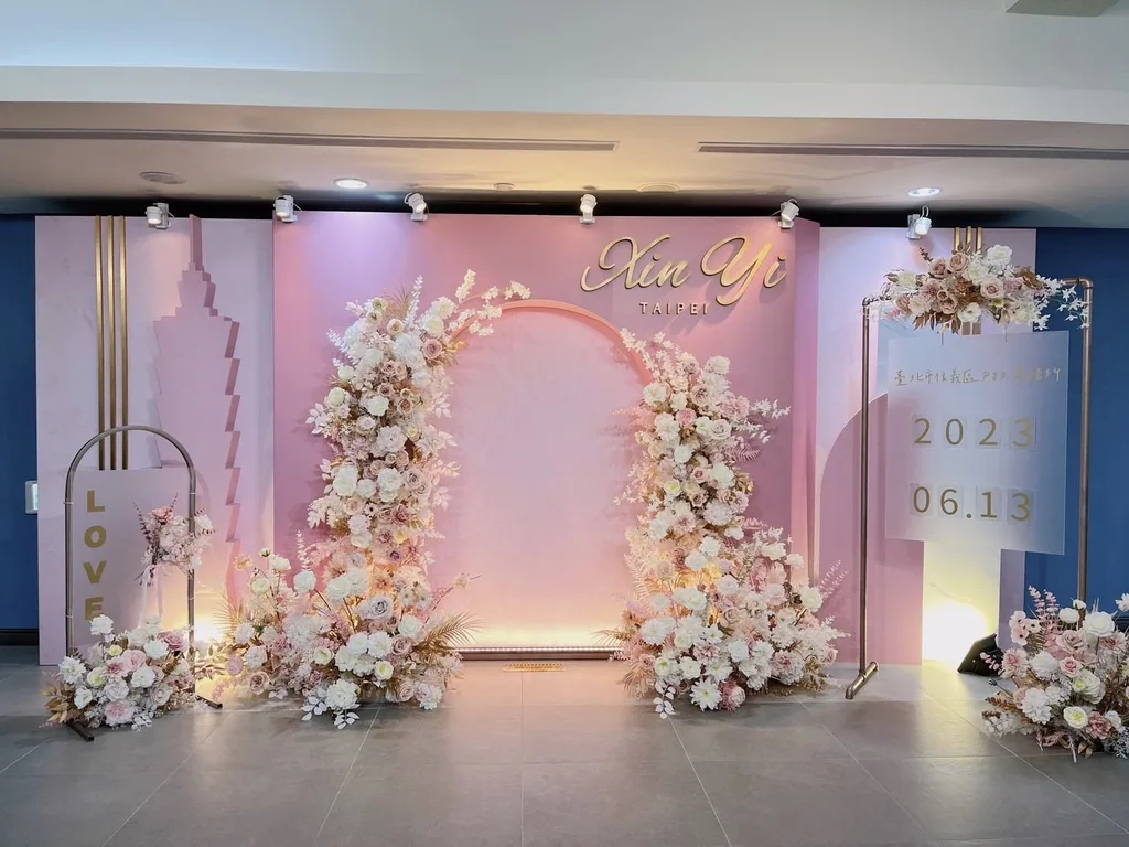 信義戶政事務所結婚登記拍照背板,2022 年 12 月施作。台北 101 做成階梯狀立體剪影立在淡粉色牆面上,中間開一個拱、兩側用米白與香檳色花材堆起花柱,右上角是金色 Xin Yi TAIPEI 立體字(畫面日期牌是拍攝當天的 2023.06.13)。