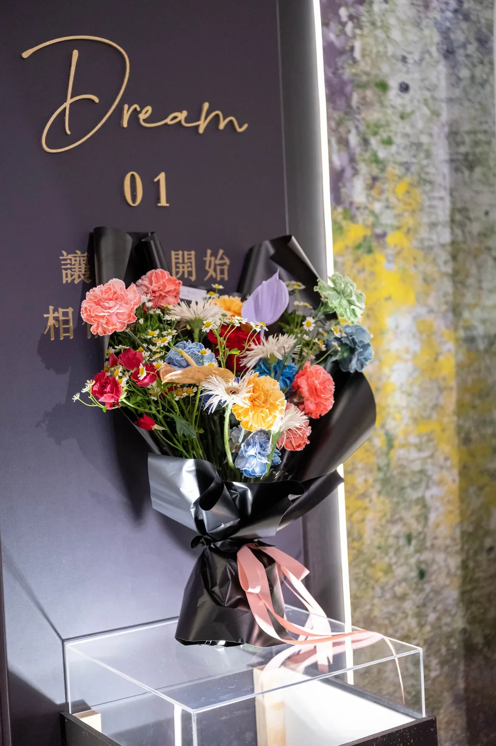 「Dream」看台前的彩色康乃馨花束，粉、紅、橘、藍、綠五色康乃馨配噴金火鶴，黑紙包裝、粉色緞帶
