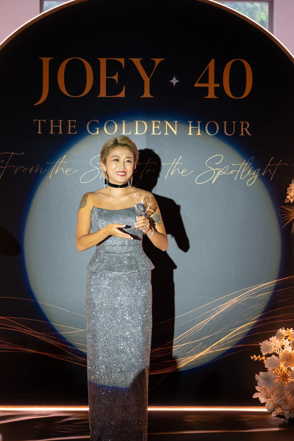 壽星 Joey 站在 JOEY 40 舞台背板前，銀色亮片禮服