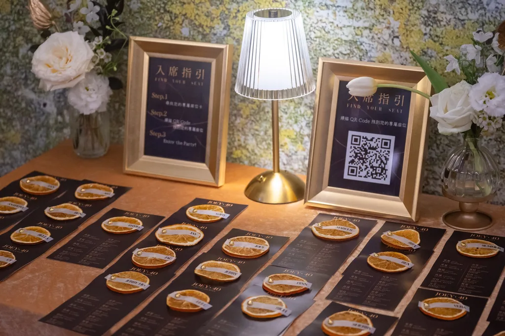 簽到桌，一排柑橘桌卡，乾橘片綁著黑底金字的專屬名卡，金框「入席指引」立牌與 QR Code，小檯燈與白色花藝