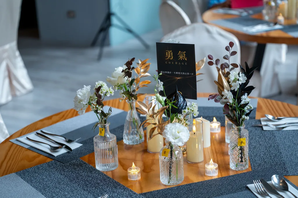 四人圓桌，幾支透明小瓶插白花與金葉、黑葉，小燭杯，桌名立牌「勇氣」立在中間
