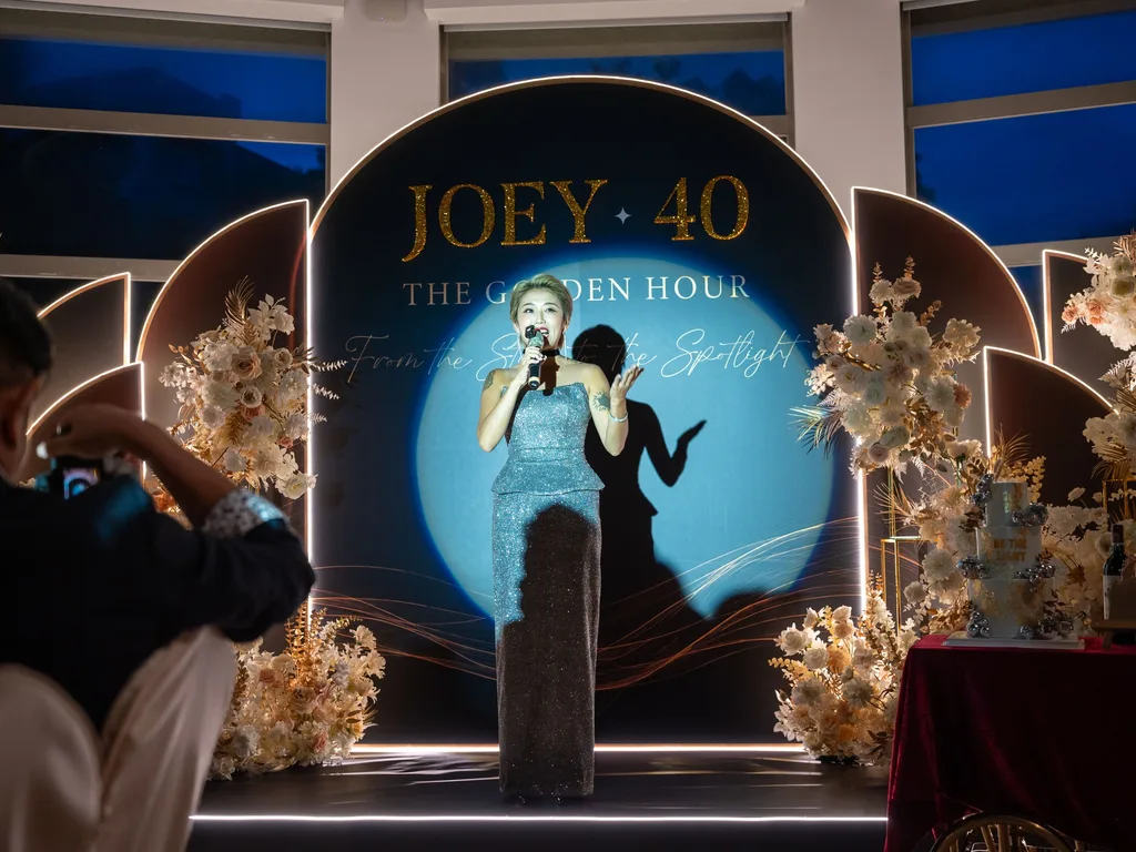 派對當晚 Joey 在舞台上致詞，聚光燈打在 JOEY 40 The Golden Hour 拱形背板上，兩側各三片 LED 邊光花瓣板與米白金色花藝，台大明達館