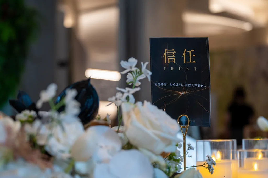 長桌上的桌名立牌「信任」，黑底金字，周圍是米白桌花與燭杯