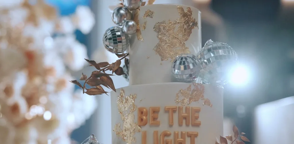 JOEY 40 生日蛋糕,白色多層蛋糕上有金箔與銀色圓球裝飾,正面寫著 BE THE LIGHT,放在紅絨桌巾的推車上