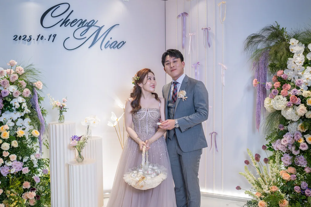新婚夫婦於淺色花卉佈置前合影,牆上婚禮日期,整體色調溫馨浪漫。