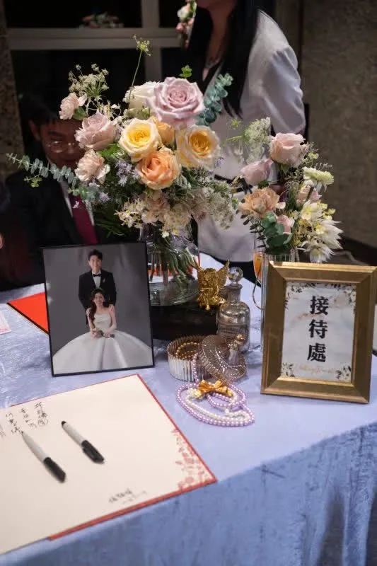婚禮接待桌,擺滿粉黃玫瑰、新人照片與簽名冊,色調溫馨浪漫。
