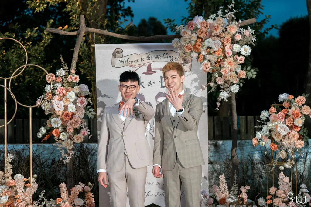 新人在美軍俱樂部戶外迎賓背板前展示婚戒,木架花藝與夜色相映成趣。