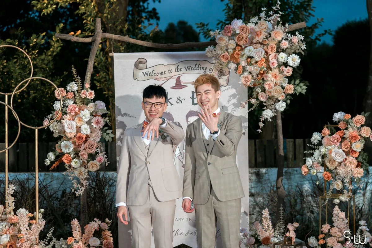 兩位新郎在戶外婚禮背景前，展示戒指，周圍是奶油色、橘粉色花藝佈置。