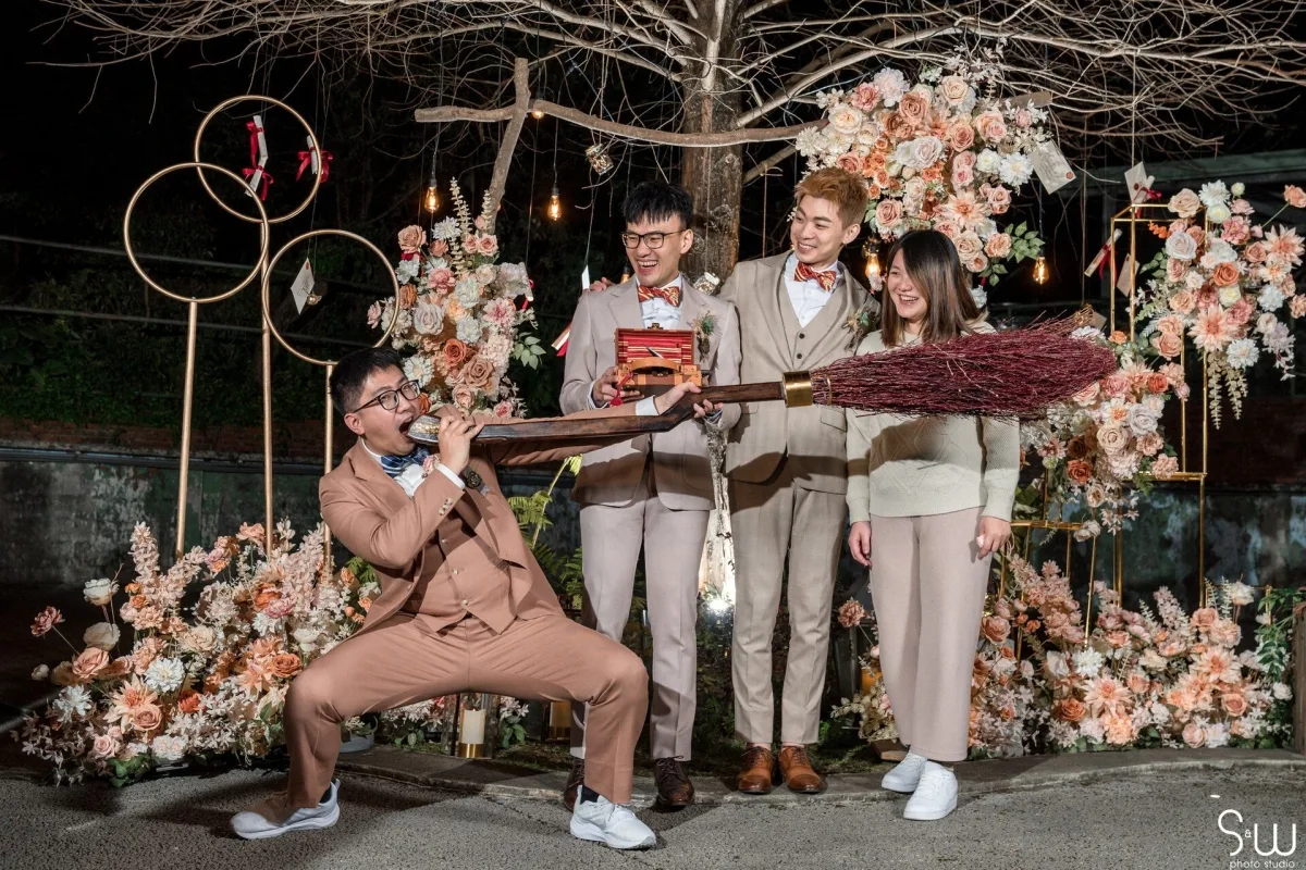 四位賓客在佈滿蜜桃、白色花朵與魔法掃帚的溫馨婚禮場景中歡樂合影。