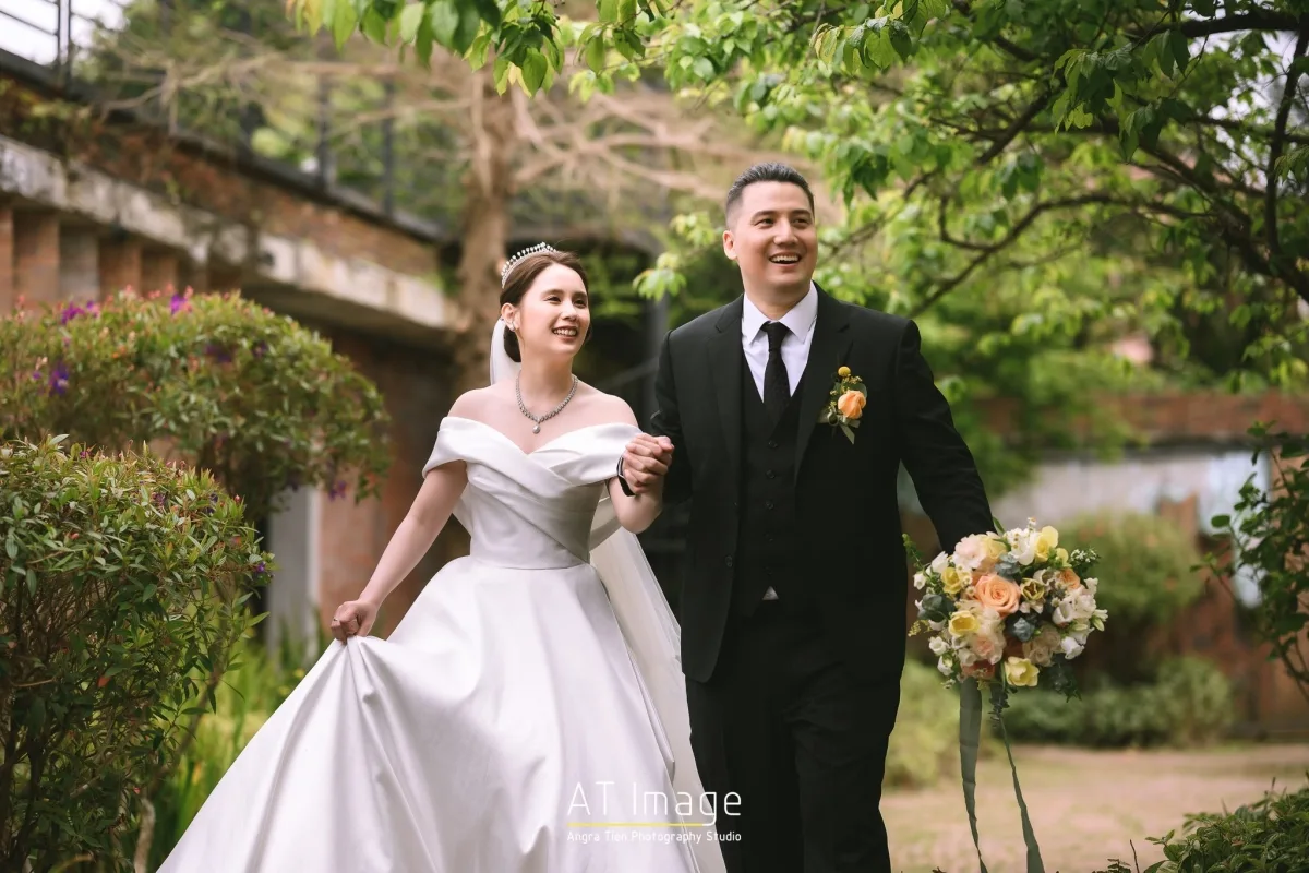 新娘新郎手牽手漫步綠意庭園，白紗黑西裝，手捧淡雅花束，色調溫馨。