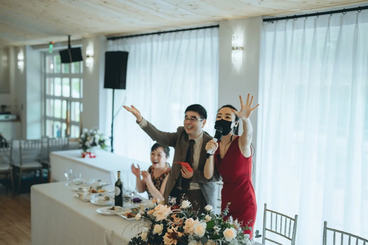 婚禮宴會，戴口罩紅裙女子持麥克風唱歌，賓客歡樂。米色花藝妝點餐桌，氣氛溫馨。