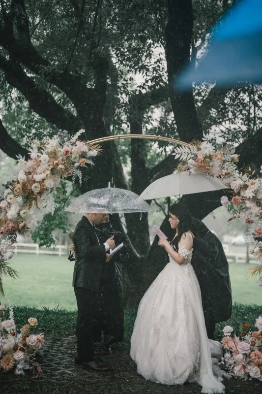 雨中婚禮，新人與證婚人撐傘，被花拱門環繞，色調柔和。