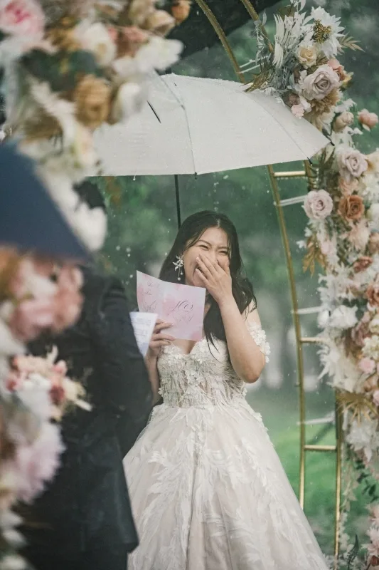 新娘在雨中婚禮上撐傘歡笑，溫和色調的花卉拱門，洋溢幸福氛圍。