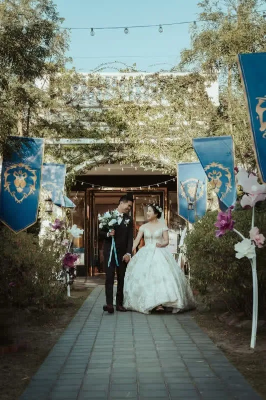 一對新人手牽手走在戶外通道,兩旁有藍旗和花飾,色調溫馨。