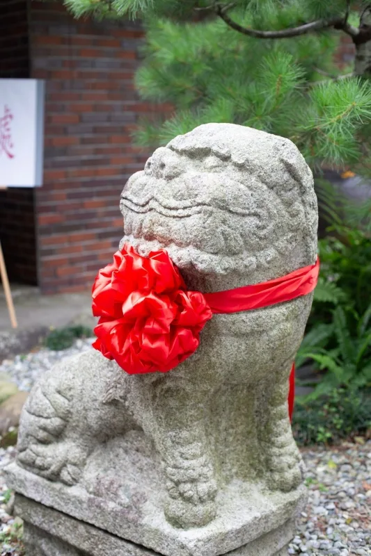 灰色石獅繫著鮮紅花結，綠色植物與紅磚背景，婚禮喜慶感十足。