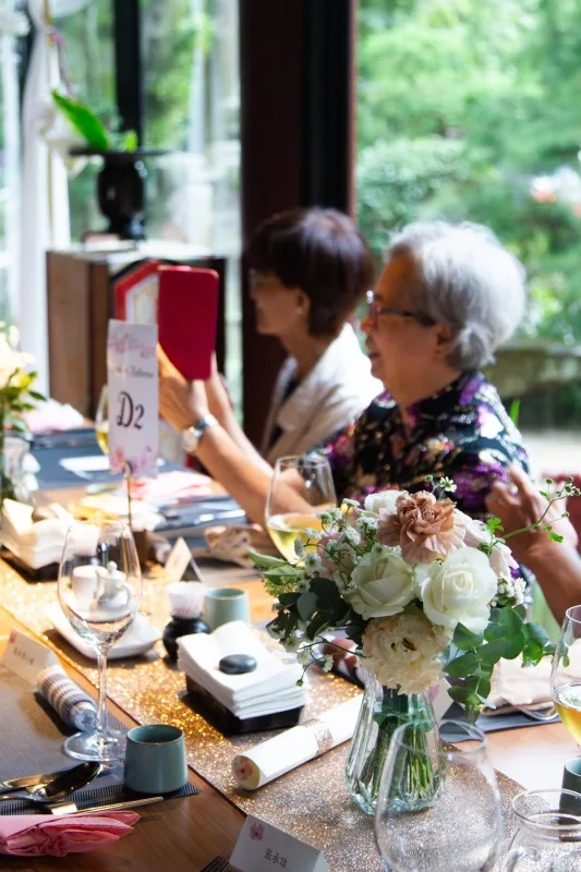 婚禮宴會餐桌佈置，有白色鮮花、金色桌旗，賓客入座，氣氛溫馨。