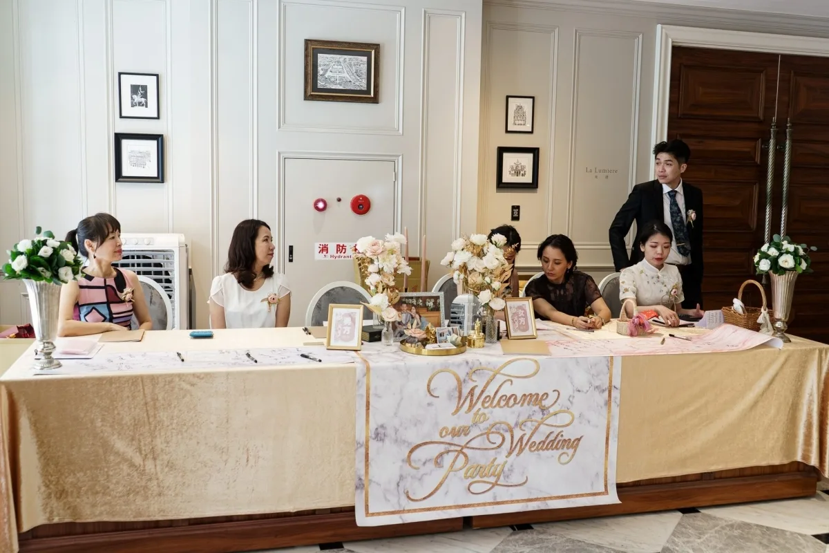 婚禮迎賓桌前，賓客簽到，金白花藝、大理石紋布條，氛圍優雅喜慶。