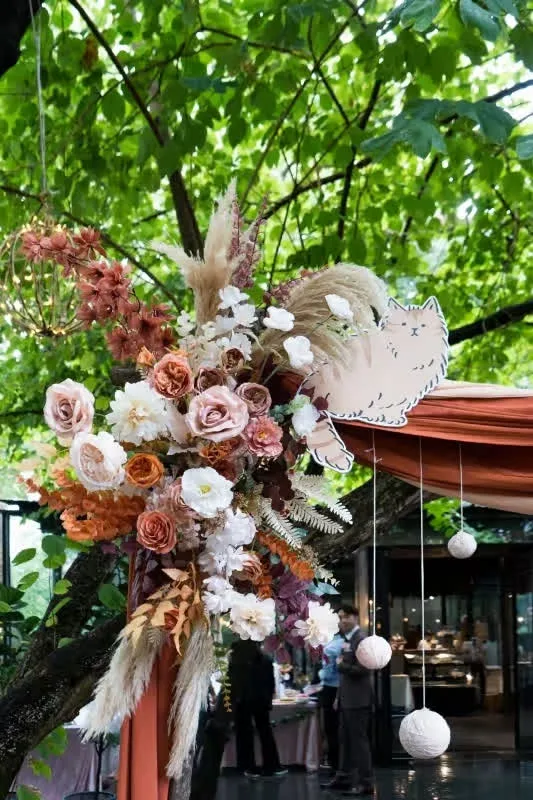粉橘色系鮮花、貓咪剪影、垂墜布幔與燈球裝飾戶外婚禮，綠樹環繞。