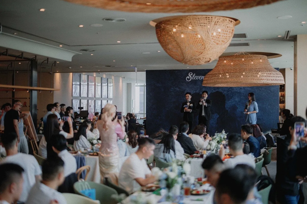 溫馨婚禮宴會，賓客圍桌而坐，台上演講者，天花板懸掛藤編燈飾，淡雅花藝點綴。