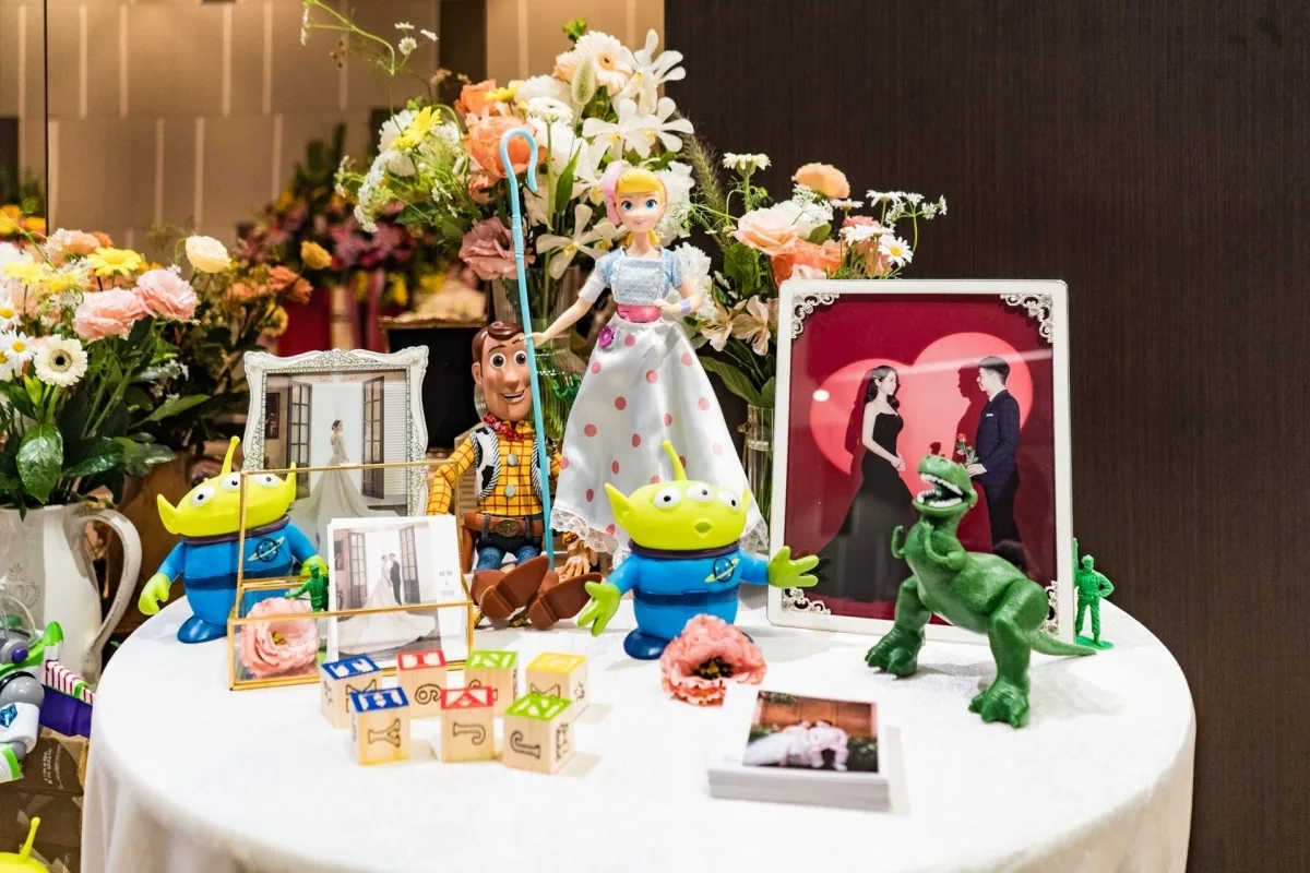 以玩具總動員為主題的婚禮擺設，胡迪、寶貝與三眼怪公仔旁，有花卉及婚紗照，色彩溫馨和諧。