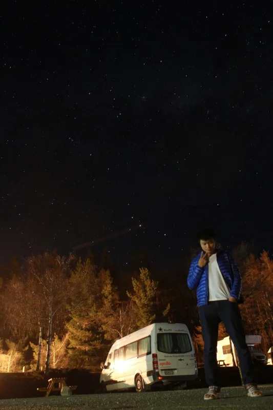 靜謐星空下，男子與露營車駐足暖光森林旁，呈現深沉夜色。