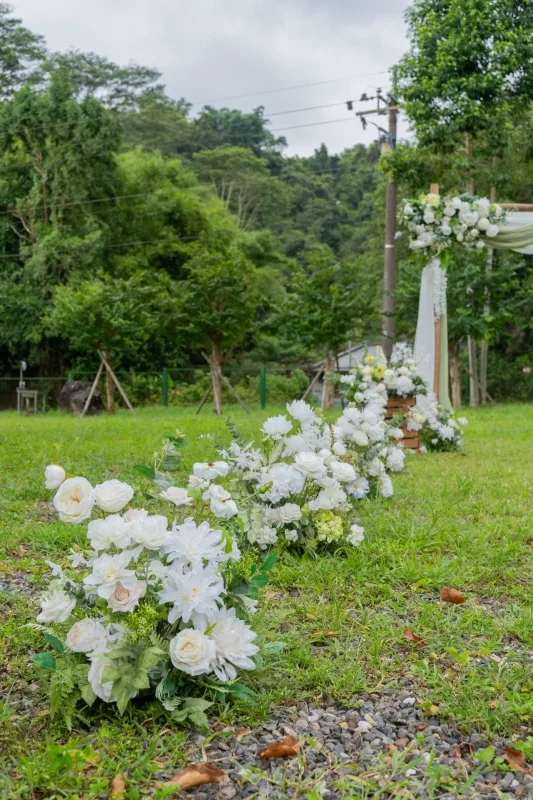 綠色草地上的婚禮走道，兩側佈滿白色花束，背景是茂密樹林，氛圍浪漫自然。