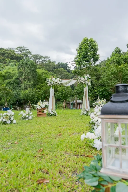 草地婚禮，白色花朵拱門、木箱與提燈佈置，背景綠樹環繞。