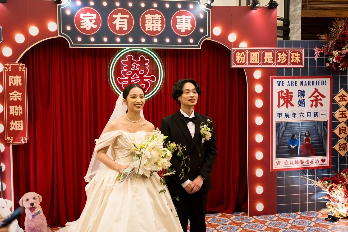 喜氣洋洋的紅色婚禮背板前，新娘穿婚紗手捧花束，新郎著西裝，甜蜜合影。