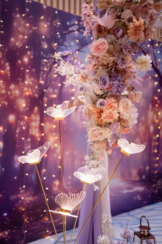 紫色夢幻背景，浪漫燈光襯托粉紫花藝與金色蝴蝶燈飾，溫馨婚禮佈置。