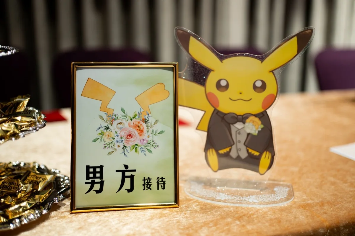 婚禮接待桌上，有金色花卉牌子與穿著燕尾服的皮卡丘立牌，暖色調。