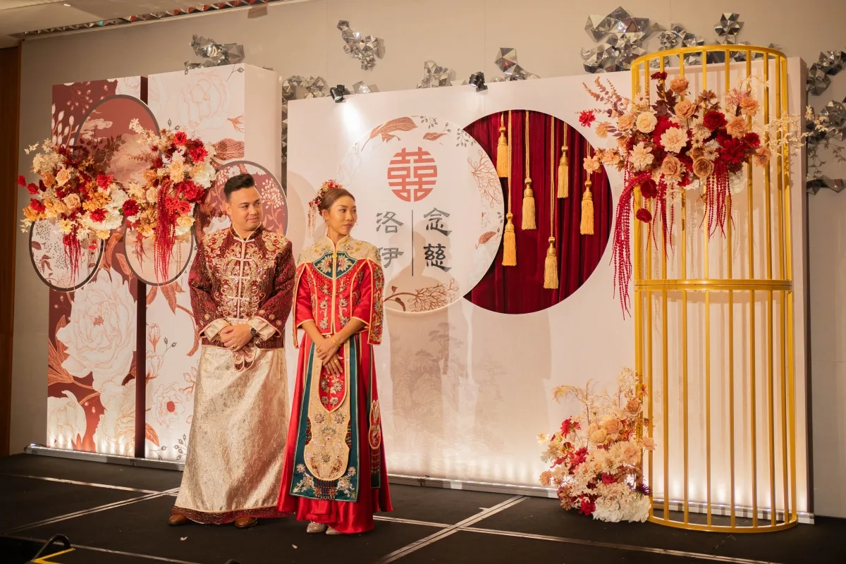 新人著中式禮服,紅金雙喜背板與花卉,喜慶傳統婚禮場景。