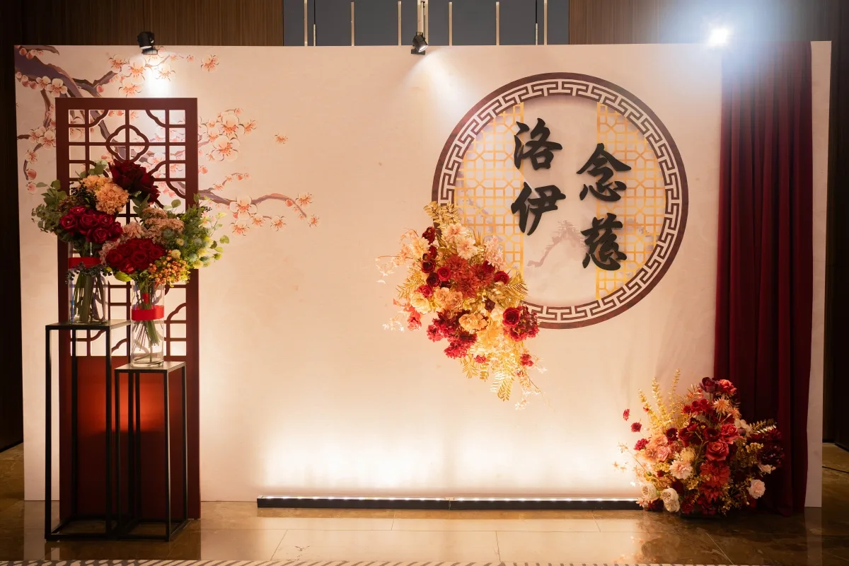 中式婚禮背景,喜慶紅金色調花藝佈置,古典窗格襯托新婚文字。