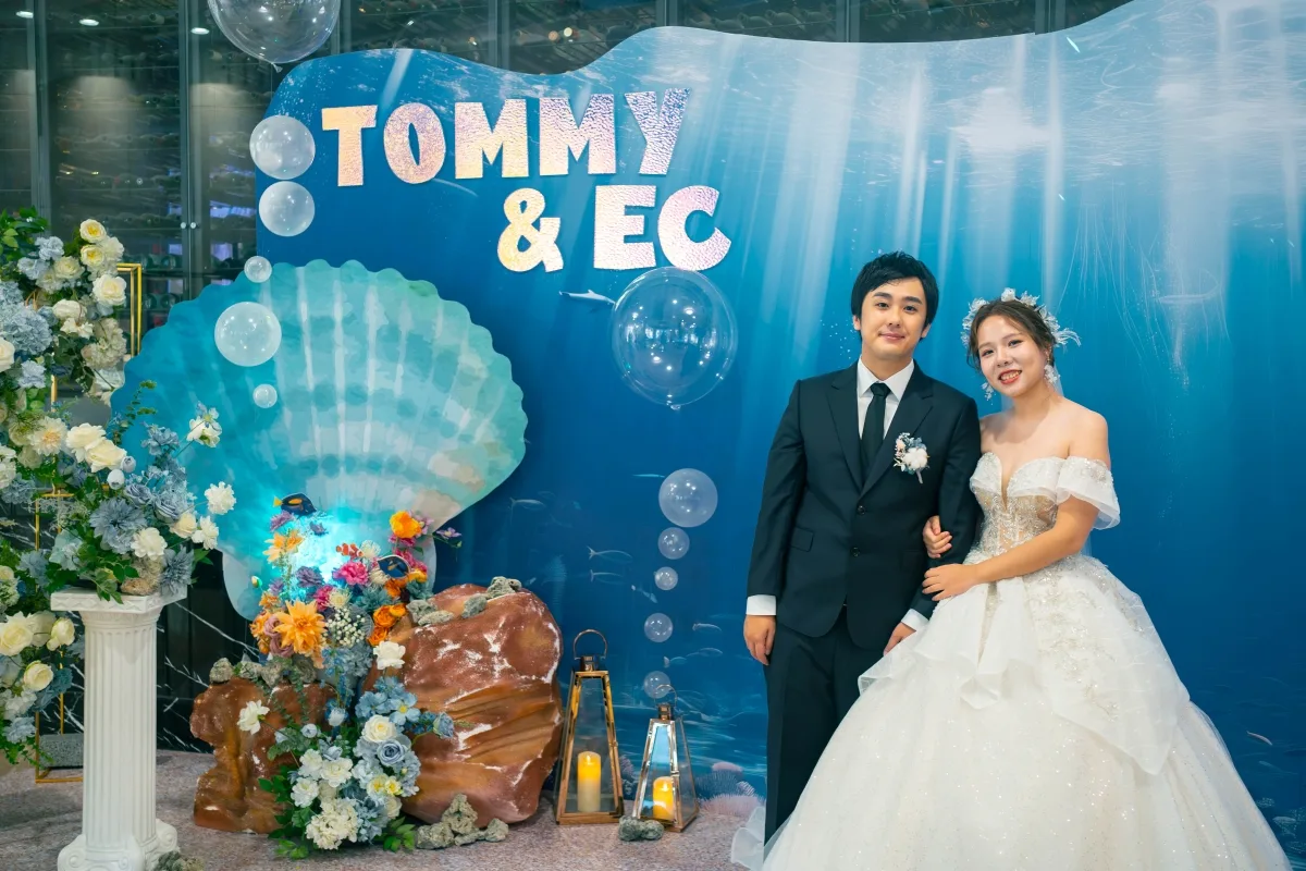 新婚夫婦站在藍色海底主題婚禮背景前,有貝殼、花卉和提燈裝飾。