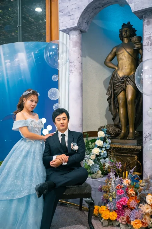 婚禮場景中,淡藍禮服新娘與黑西裝新郎依偎,藍白花卉雕像環繞,充滿浪漫夢幻氣息。