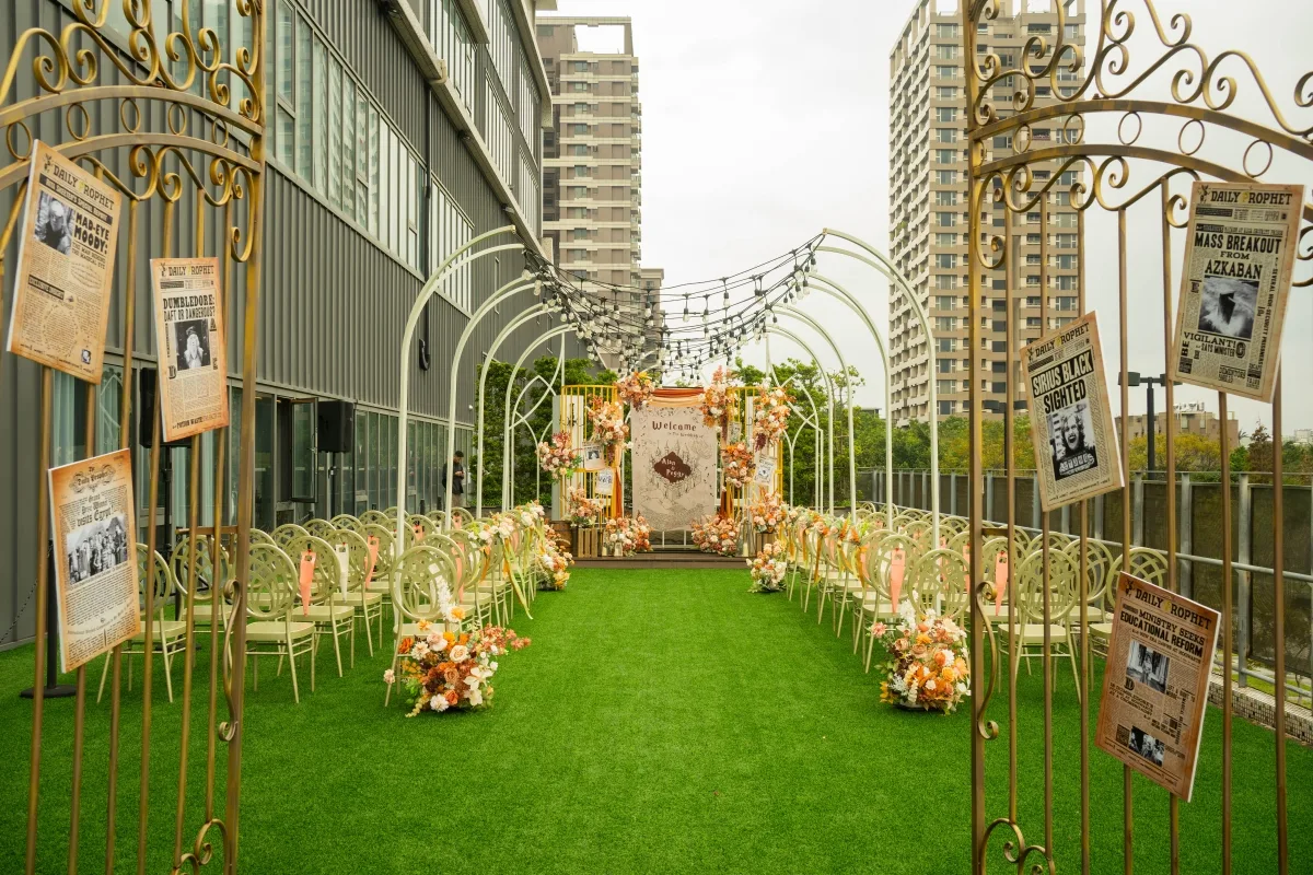 戶外婚禮場地，綠草地毯、金白拱門、暖色花藝、燈串及報紙裝飾。