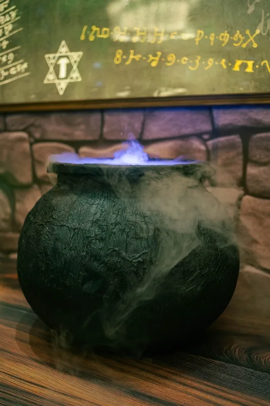 冒著藍光的黑色巫婆鍋，煙霧繚繞，後方為神秘符號與磚牆背景。