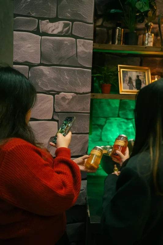 兩位賓客手持特調飲料,一人手機拍攝,背後有婚紗照與綠色燈光點綴。