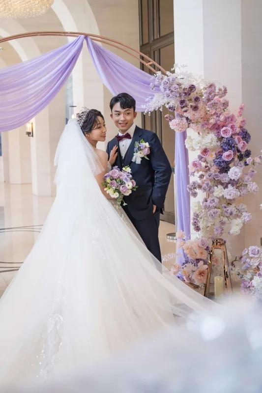 浪漫婚禮，一對新人在紫色、粉色花拱門前合影，新娘手持紫色捧花。