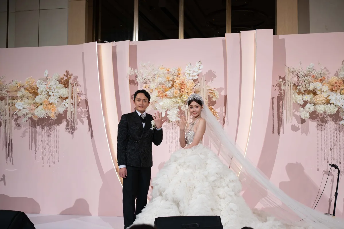 婚禮上，新婚夫婦身穿禮服於粉色花牆前，展示幸福婚戒。
