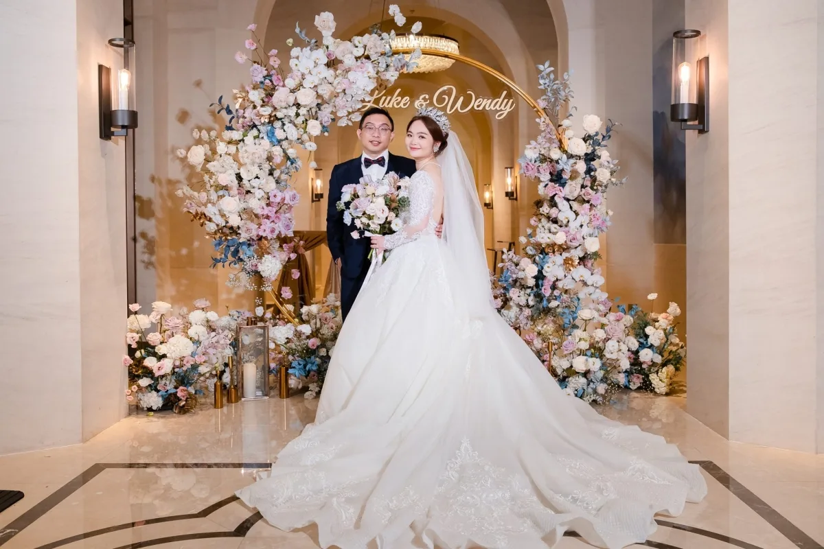 新婚夫婦在金色花卉拱門前合影，淡雅粉藍色調，浪漫幸福。