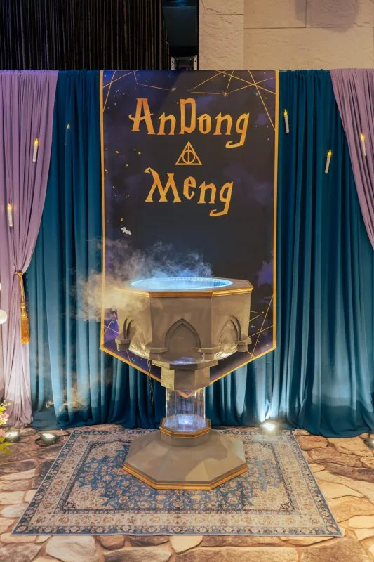 魔法主題婚禮場景，湛藍與薰衣草色帷幕襯托冒煙坩堝，金字旗幟上書新人名字。