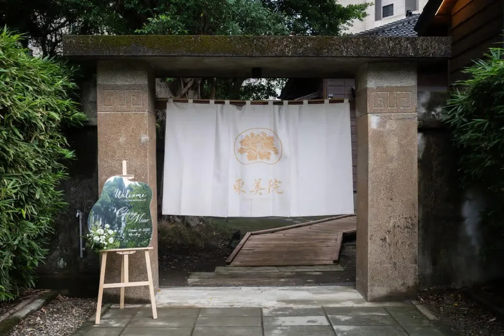 古樸石門前，白色布幔上金花圖騰與「東美院」字樣，迎賓牌旁點綴白花，婚禮氛圍靜謐雅致。