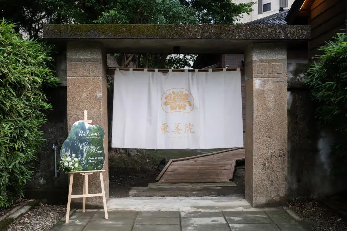 古樸石門前，白色布幔上金花圖騰與「東美院」字樣，迎賓牌旁點綴白花，婚禮氛圍靜謐雅致。