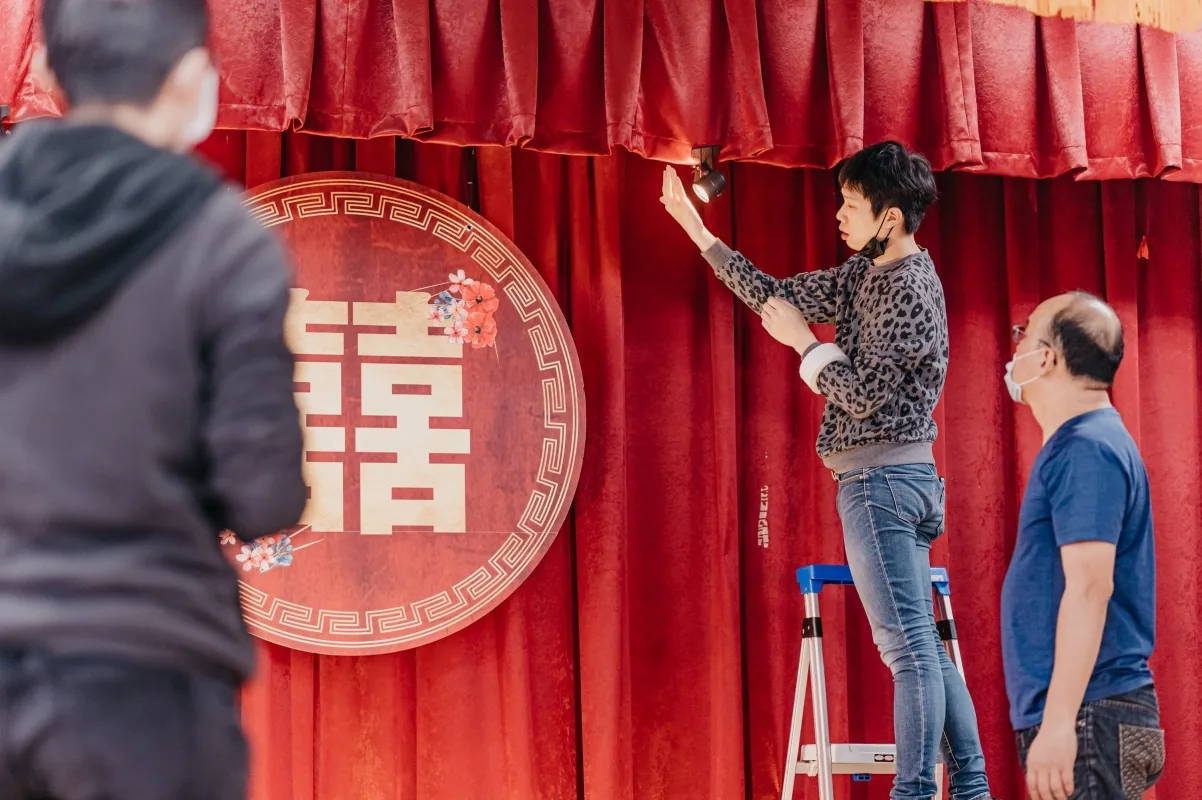 工作人員在紅色背景前，佈置著金色雙喜字婚禮裝飾，營造喜慶氛圍。