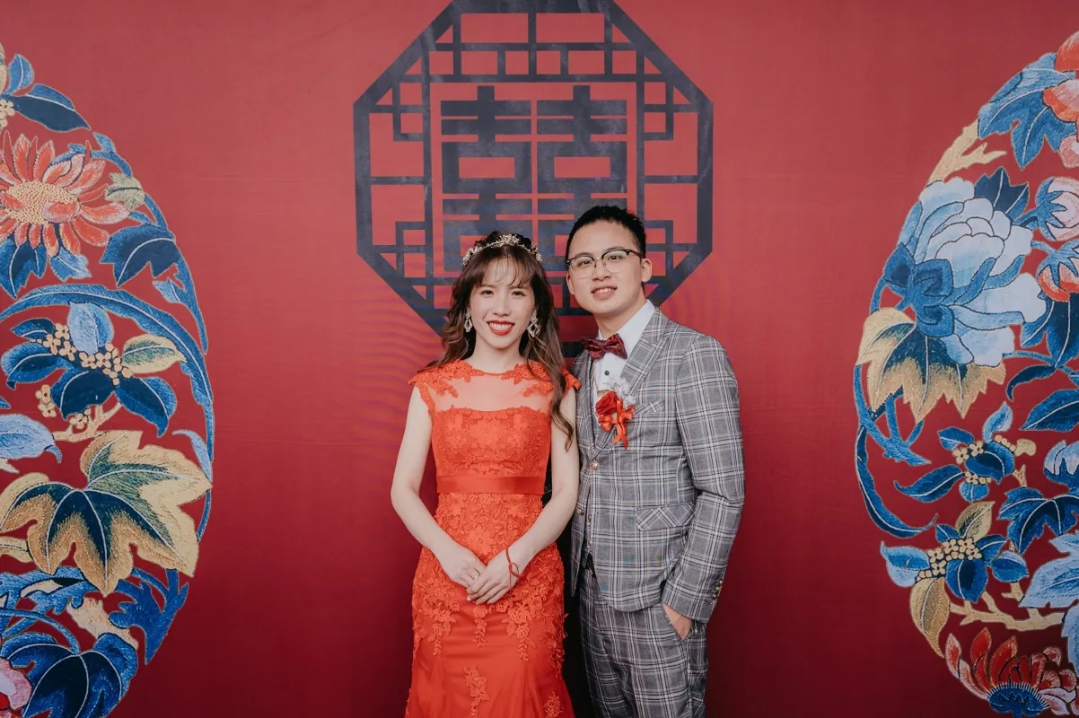 紅色背景中，一對新婚夫婦穿著禮服，背景有雙喜字與花卉圖案，洋溢喜氣。